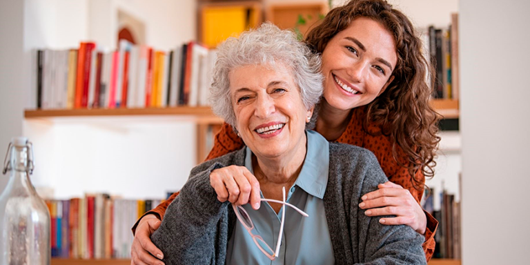 Lächelnde pflegende Angehörige umarmt ältere Frau im heimischen Wohnzimmer – ein Bild, das die enge Beziehung und die Bedeutung von Pausen in der häuslichen Pflege zeigt. Die IAHA (International Association for Healthy Aging) unterstützt pflegende Angehörige in der Schweiz mit Beratung, Schulungen und praktischen Tipps für mehr Entlastung und Lebensqualität