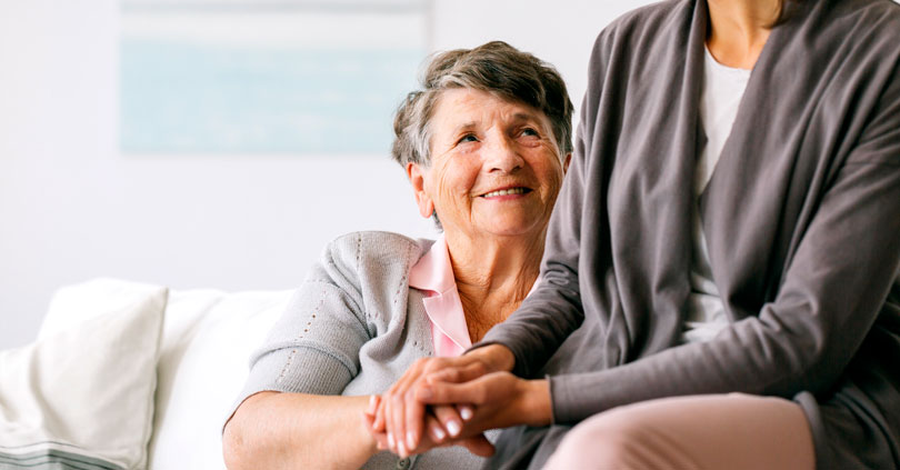 Eine ältere Frau sitzt lächelnd auf einem Sofa und hält die Hand einer jüngeren Betreuungsperson, die fürsorglich neben ihr sitzt. Die Szene veranschaulicht einfühlsame Demenzpflege im Alltag. Die IAHA (International Association for Healthy Aging) unterstützt pflegende Angehörige mit praxisnaher Beratung, Schulungen und Entlastungsangeboten.